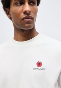 Άνδρας με λευκό φούτερ που έχει μικρό κόκκινο μήλο και το κείμενο "φρεσκάδα σε μια γουλιά marc o'polo denim" στο στήθος, κοιτάζει πλάγια.