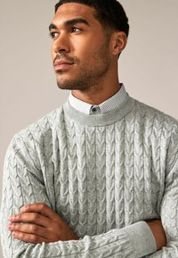 Un pull en tricot cable gris clair avec un col haut, porté sur une chemise à carreaux. Manches à bords côtes visibles et détails de texture complexes.