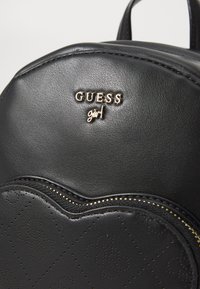 Sac à dos en cuir noir avec un logo doré "GUESS girl", une texture lisse et une poche zippée avec un motif perforé.