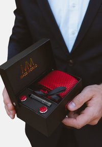 Scatola regalo nera che mostra una cravatta rossa testurizzata, gemelli e una penna. Include il logo dorato "MASSI MORINO" all'interno della scatola. Nastro decorativo incluso.