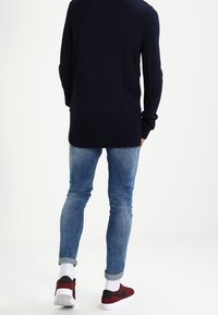 Pull en maille à manches longues bleu marine, jeans skinny bleu clair, et baskets rouges et noires avec semelles blanches, présentant des poignets retroussés et une texture lisse.
