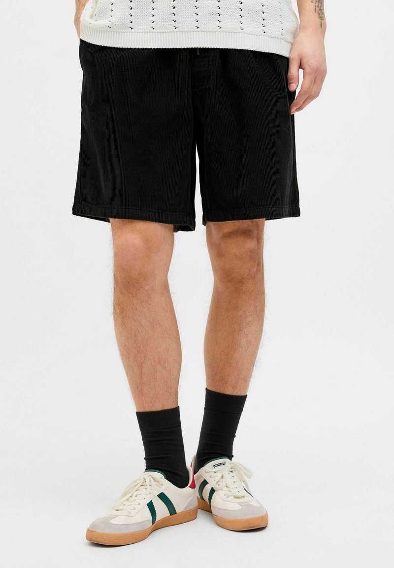 Sorte fløjlsshorts med en afslappet pasform. Har brede ben og et struktureret stof. Sammen med hvide sneakers og sorte sokker.