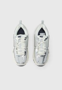 Boty Nike s stříbrným síťovaným svrškem, bílými doplňky a polstrovanou podrážkou. Mají kulaté tkaničky a poutko pro snadné obouvání.