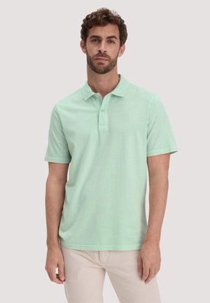 Man in een lichtgroen poloshirt met korte mouwen en lichtbeige broek, staand tegen een effen lichtgrijze achtergrond.