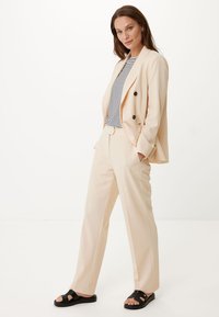 Beige skräddarsydd kostym med dubbelknäppt kavaj och raka byxor, kombinerad med en randig skjorta och svarta sandaler.