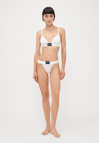 White bikini set featuring a triangular bralette with thin straps and a matching thong bottom. Both pieces have a logo band. Smooth fabric.