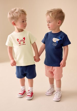 Deux jeunes garçons se tenant la main, portant des shorts et des t-shirts avec des motifs d'animaux marins, debout sur un fond neutre.