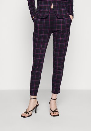 Mujer vestida con pantalones a medida de cuadros azul marino y púrpura y blazer a juego, combinados con sandalias negras de tacón alto con tiras.