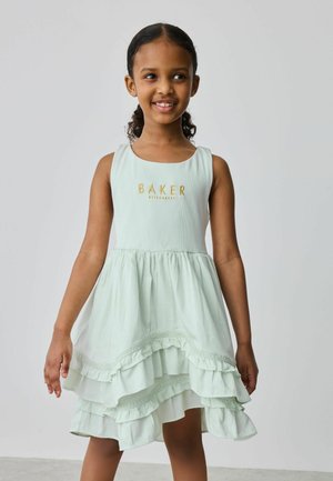 Junges Mädchen lächelt, trägt ein ärmelloses, blassgrünes, mehrlagiges Kleid mit dem Schriftzug "BAKER" auf dem Oberteil und steht vor einem schlichten hellgrauen Hintergrund.