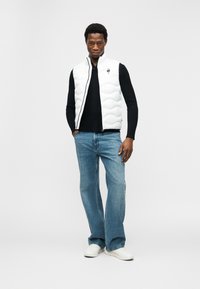 Άνδρας όρθιος με τα χέρια στις τσέπες, φοράει λευκό καπιτονέ γιλέκο πάνω από μαύρο πουκάμισο, μπλε τζιν και λευκά αθλητικά παπούτσια σε απλό φόντο.