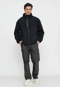 Giacca nera con zip e parte superiore testurizzata, dettaglio del logo e cappuccio, abbinata a pantaloni cargo grigio scuro e sneaker chiare.