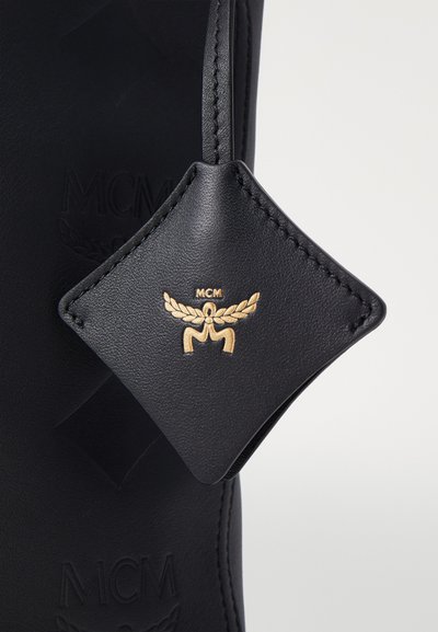 Mustast nahkasilmal on kuldne logo, mis on kinnitatud õhukese rihmaga, ning sellel on läikiv kuju ja õmblused. Taustal on joonistatud kaubamärk.