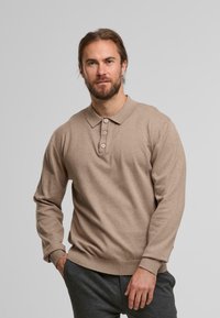 Man met baard in een beige langarmige polo shirt en donkergrijze broek, staand met één hand in zijn zak tegen een effen achtergrond.