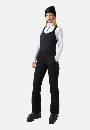 Woman wearing black ski bib pants, white long-sleeve zip-up top, black beanie, and black ski boots, standing with hands in pockets.