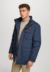 Giacca puffer blu navy con chiusura a zip, numerose tasche e colletto rialzato. Presenta dettagli cuciti e un logo sul petto.