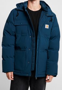 Hombre vestido con una chaqueta acolchada azul con capucha, cremallera y botones a presión, bolsillos de solapa en el pecho y parche de logo en el lado izquierdo.