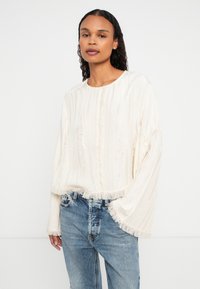 Eine Frau mit langen lockigen Haaren trägt eine lockere cremefarbene Bluse mit fransigen Rändern und hellblauen Jeans und steht vor einem schlichten weißen Hintergrund.