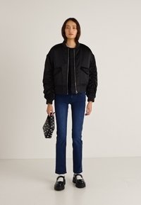 Chaqueta bomber negra con mangas abullonadas, dos bolsillos delanteros, combinada con jeans azul oscuro y zapatos negros, sosteniendo un bolso de lunares.