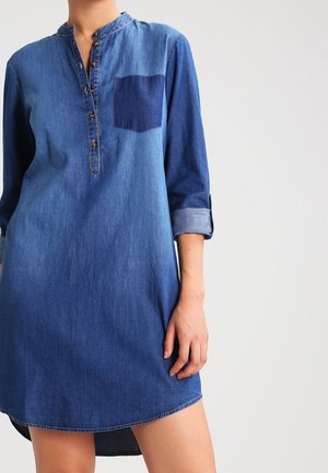 Woman wearing a casual blue denim shirt dress with three-quarter sleeves and a front pocket, standing against a plain light background.