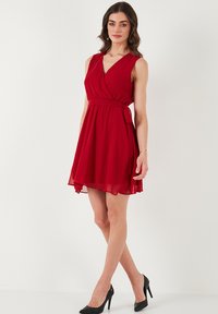 Red, sleeveless wrap dress with a V-neck, cinched waist, and flowing skirt. Made of lightweight fabric with a soft texture. Black high heels.
