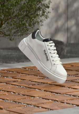 Biały sneaker marki Valentino z czarnym panelem na pięcie unosi się nad drewnianymi płytkami na zewnątrz, w tle zielona roślina i szara ściana.