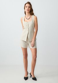 Linnenmix outfit met verticale strepen, bestaande uit een fitted gilet met knoopsluiting en bijpassende shorts; gecombineerd met zwarte puntige hakken.