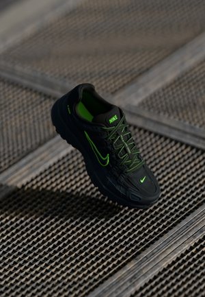 Czarny sneaker Nike z neonowozielonymi detalami i sznurowadłami, unoszący się nad teksturowaną metalową kratą.