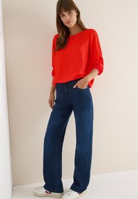 Rotes Langarmshirt, kombiniert mit hochtaillierten, weit geschnittenen dunkelblauen Jeans und weißen Turnschuhen mit Farbakzenten. Glatter Stoff, lockere Passform.