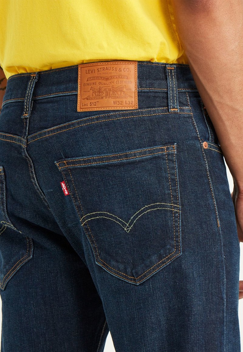 Un jean en denim bleu foncé présente une patch en cuir marron portant l’inscription « Levi Strauss & Co. » avec des coutures jaunes et un design de poche arrière courbée emblématique.