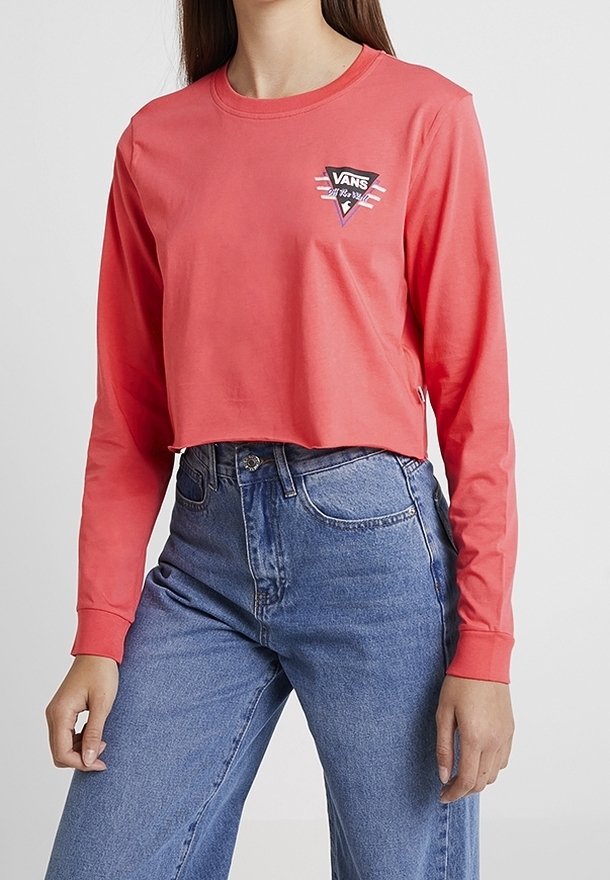 Korallefarbene, langärmlige, gekürzte Bluse mit rundem Halsausschnitt und Vans-Logo; kombiniert mit hoch taillierten blauen Jeans. Weiches Baumwollmaterial.