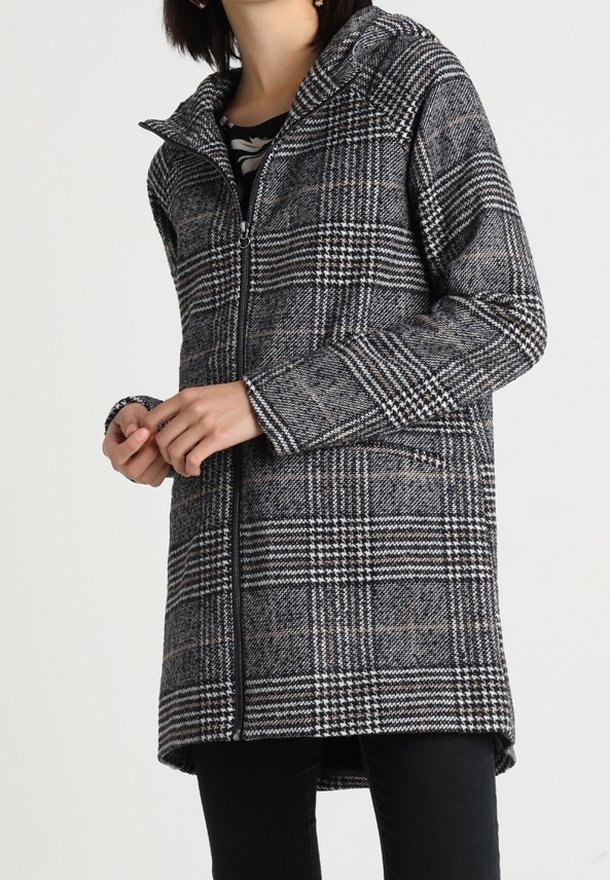 Femme portant un manteau zippé à carreaux gris et noir avec un col montant, debout les mains jointes, sur un fond blanc uni.