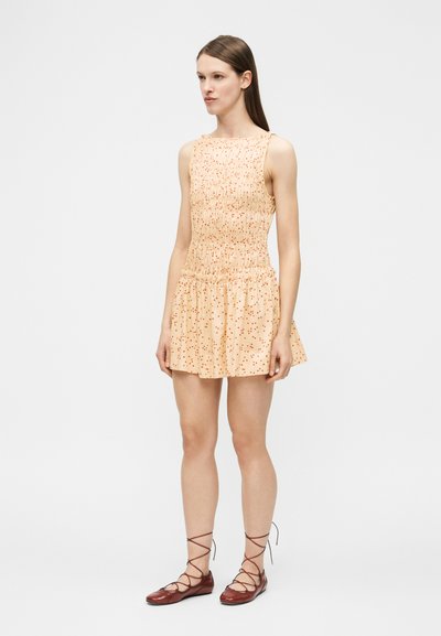 Γυναίκα που στέκεται φορώντας ένα αμάνικο ροδακινί φόρεμα με κόκκινες πουά και καφέ δερμάτινα επίπεδα παπούτσια με κορδόνια, σε έναν απλό λευκό φόντο.