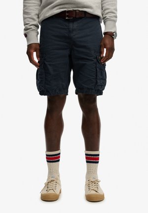 Mężczyzna noszący granatowe szorty typu cargo, beżowe tenisówki, kremowe skarpetki z czerwonymi i granatowymi paskami, szarą bluzę, brązowy skórzany pasek i zegarek na rękę.