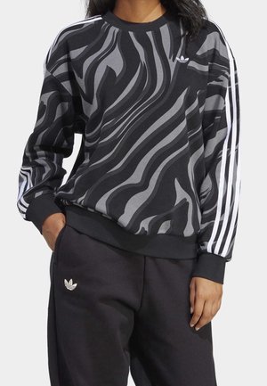 Osoba ubrana w czarną i szarą bluzę z wzorem wirowym, z białymi paskami na rękawach oraz czarne spodnie Adidas z logo na kieszeni.