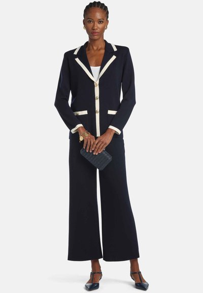 Navy blazer with cream trim and gold buttons, paired with wide-leg pants. The model holds a textured dark clutch and wears navy shoes.