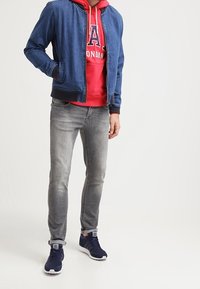 Blouson bomber en denim bleu foncé porté sur un sweat à capuche rouge avec texte imprimé. Jean gris slim et baskets navy avec des accents blancs.
