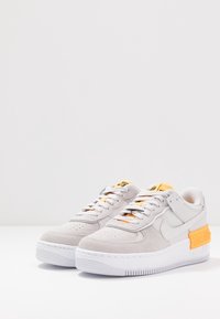 Šedé semišové a kožené tenisky s bílými podrážkami, oranžovými akcenty a černými tkaničkami. Kulatá špička a polstrovaný límec pro pohodlí.