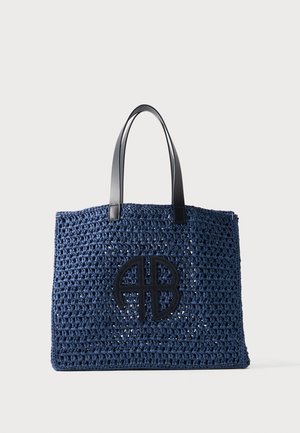 Sac cabas bleu marine crocheté avec des poignées en cuir noir et un logo circulaire noir présentant les lettres A et B sur le devant.