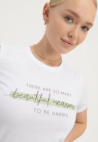 Hvid bomuld t-shirt med trykt tekst "Der er så mange smukke grunde til at være glad," med grønne accenter og flydende skriftdesign.