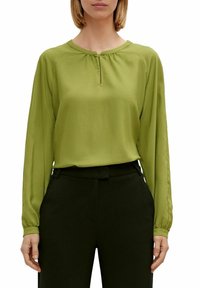 Blusa verde a maniche lunghe con scollo rotondo e piccolo dettaglio a chiave frontale, realizzata in tessuto morbido, abbinata a pantaloni neri con texture.