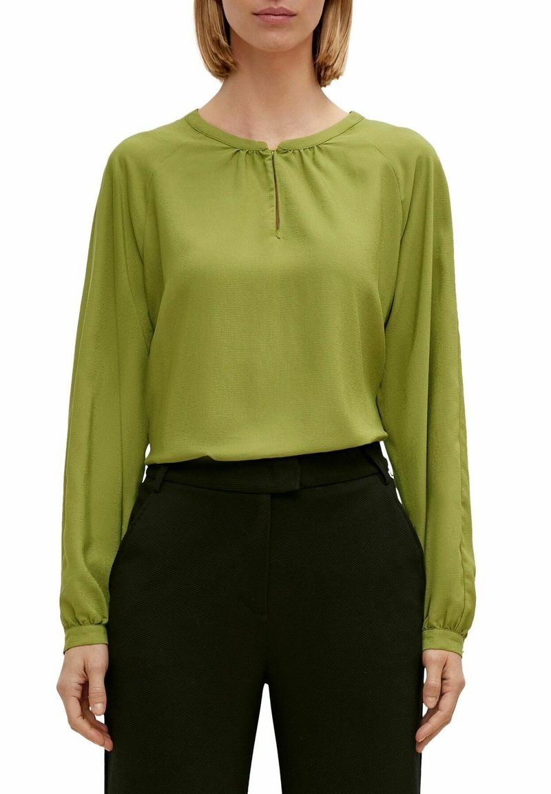 Blusa verde a maniche lunghe con scollo rotondo e piccolo dettaglio a chiave frontale, realizzata in tessuto morbido, abbinata a pantaloni neri con texture.