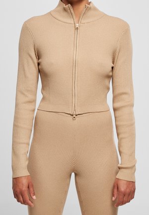 Person wearing beige ribbed knit zip-up long sleeve top and matching pants against plain background.