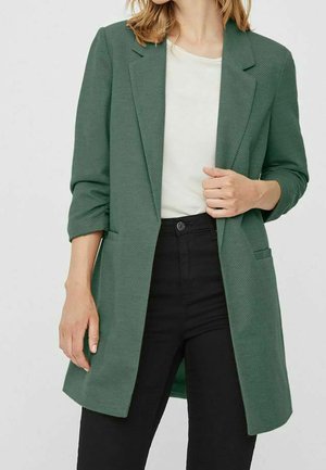 Mujer con blazer verde oscuro con mangas remangadas, camisa blanca y pantalones negros de talle alto frente a un fondo liso.