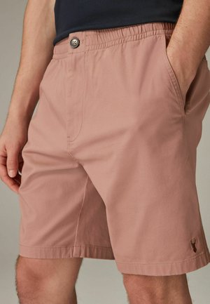 Mand iført støvet lyserøde shorts med sidelommer, knaplukning, elastisk talje og lille syet hjertelogo på venstre kant.