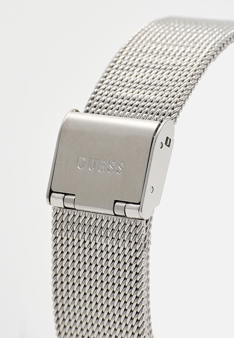 Correa de reloj de malla metálica plateada con un cierre cepillado que presenta el nombre de la marca "GUESS" grabado sobre un fondo blanco.