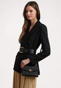 Sac à main en cuir texturé noir avec un logo doré. Dispose d'une bandoulière en chaîne, d'un petit rabat et d'une forme structurée pour un transport facile.
