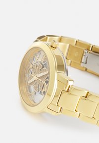 Gold metal wristwatch with a round face, skeleton design, glitter accents, and a polished bracelet strap featuring a clasp closure.