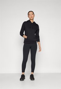 Svart huvtröja med framficka och logotyp, i kombination med svarta leggings med sidfickor och svarta träningsskor.