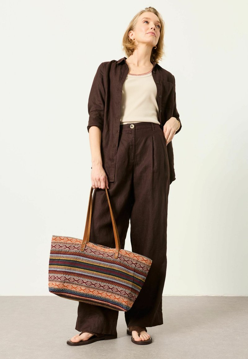 Woman wearing dark brown wide-leg pants, matching shirt over beige tank top, holding a large multicolored patterned tote bag with leather straps.