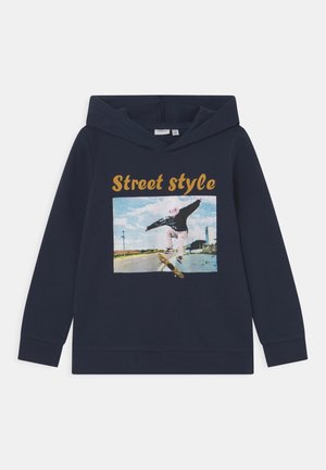 Sweat à capuche bleu marine avec un skateur réalisant un flip sur une route vide sous un ciel bleu, texte "Street style" au-dessus du graphique.